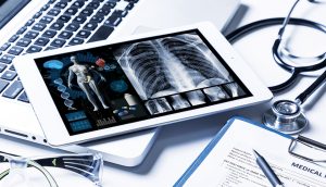 Radiology imaging on tablet screen with laptop, paper medical chart, goggles, and stethoscope.